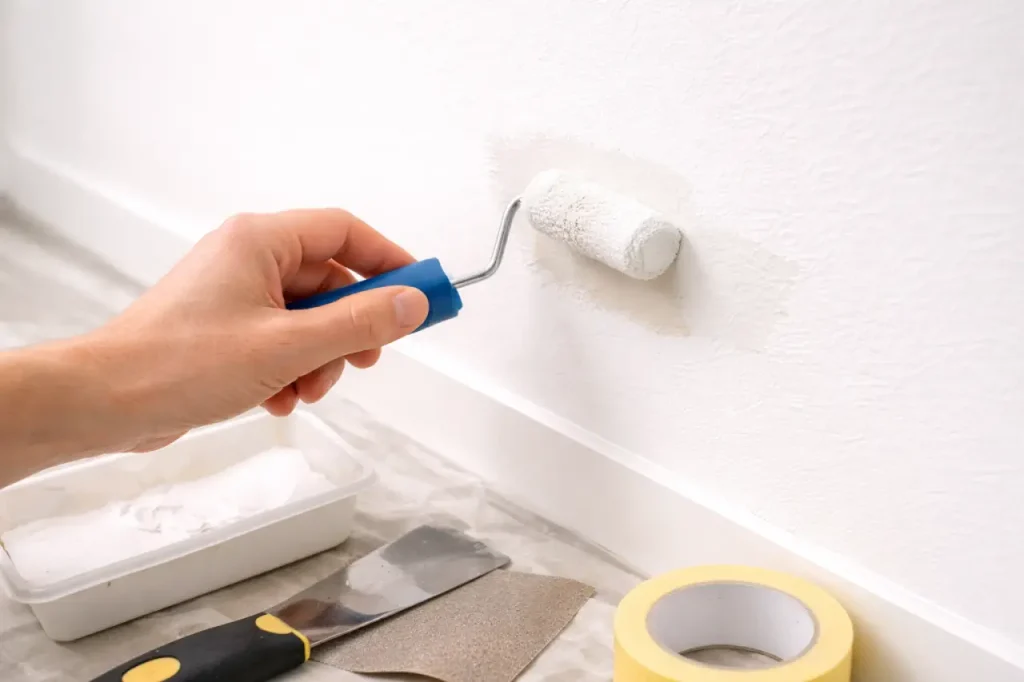 Main appliquant de la peinture au mini-rouleau pour réparer une zone arrachée sur un mur blanc, avec matériel de masquage au sol